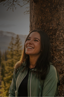A woman smiling and looking upward while leaning against a large tree trunk outdoors, with mountains and evergreen trees in the background. She is wearing a light olive green zip-front jacket over a black top.