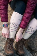 A close-up lifestyle shot of a person sitting on rocks, tying the laces of black hiking boots while wearing beige patterned compression leg sleeves and a burgundy puffer jacket. - product view 3